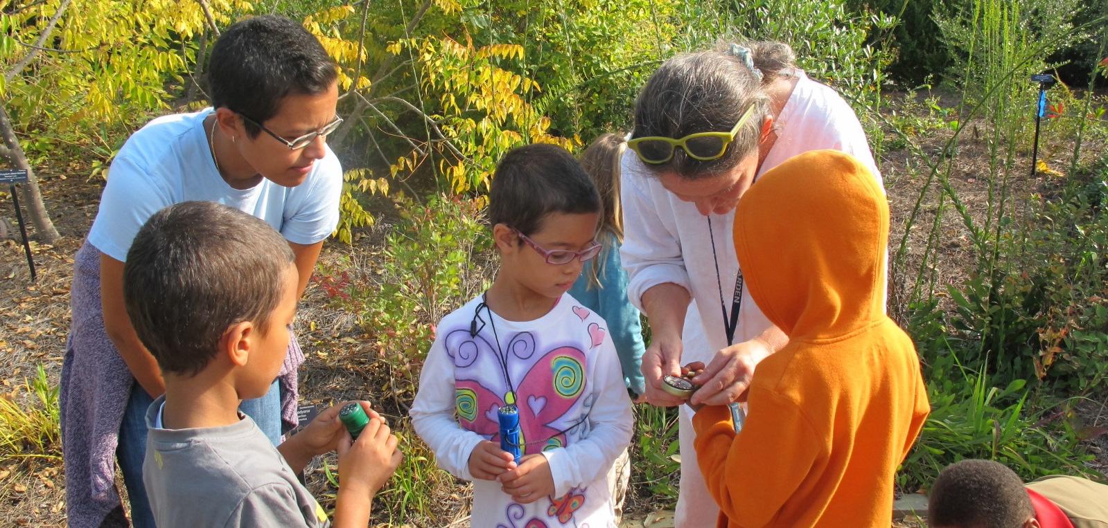 three young kids view a hand lens held by an instructor in an outdoor garden